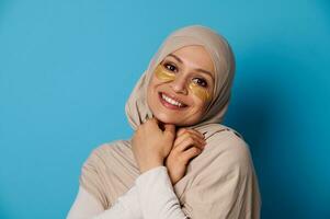 Beautiful woman with covered head in hijab, smiling at camera with hydrogel collagen patches under eyes photo