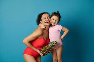 un linda pequeño niña abrazando su madre con un piña en su brazos, ambos en baños trajes. contento madres día y verano conceptos en azul antecedentes. foto