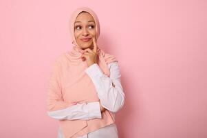 Adorable young Muslim gorgeous beautiful woman with a covered head in hijab looks mysteriously at a pink background with copy space and puts her finger to the corner of her lips, smiling thoughtfully photo