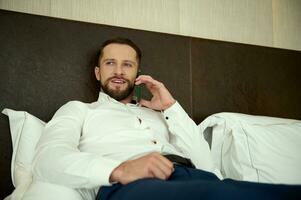 Charming attractive middle aged European successful businessman, smiles talking on mobile phone, lying on a bed in the bedchamber of a hotel, relaxing after hard day at work and business meetings photo