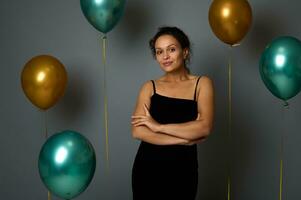 Confident portrait of gorgeous elegant Latin American woman in black evening dress posing with crossed arms looking at camera against gray background with gold and green metallic air balloons photo