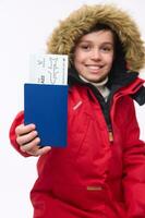 Focus on a boarding pass and passport in the hand of a blurred adorable preteen, elementary aged boy in bright red parka, smiling toothy smile looking at camera. Winter holidays travel concept photo