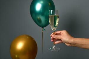 Close-up of female hand holding a glass with sparkling wine, isolated over gray wall background with inflated beautiful golden and green air balloons. Christmas, New Year and Anniversary concept photo