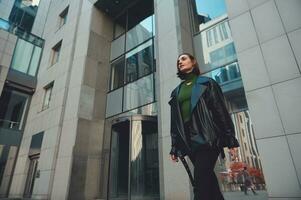 de moda morena mujer con elegante corto Corte de pelo caminando a lo largo el calle con moderno corporativo Alto edificios en el antecedentes foto