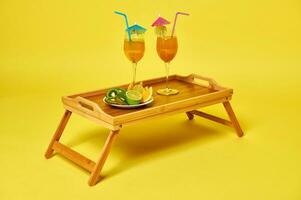 Two glasses with exotic summer cocktails decorated with cocktail umbrella and plate with sliced fruits on bamboo serving tray photo