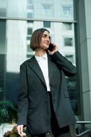 Business and communications concept. Beautiful self-confident woman, freelancer, entrepreneur, copywriter talking on cell phone on modern office corporate high-rise buildings background. photo