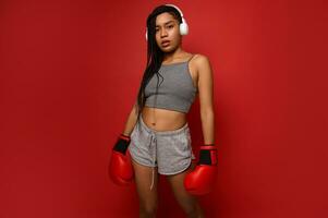 Young sporty African American woman boxer in wireless headphones and sportswear poses with boxing gloves against red background with copy space. Contact martial art concept photo