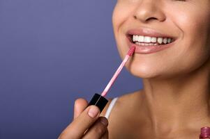 Close-up. Cropped view of attractive smiling woman with perfect clean healthy skin applying lip gloss hygienic balm on plump lips isolated on purple background. Copy ad space. Make-up concept photo