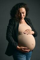 latín americano Rizado confidente embarazada madre conmovedor su embarazada barriga en el embarazo trimestre tercero, gris antecedentes foto