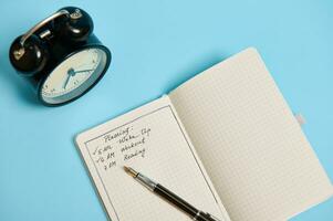 High angle view of an open organizer with plans for the day, ink pen and alarm clock on blue background with copy space. Time management, business and planning concept photo