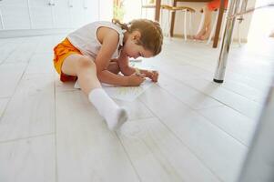 Adorable little kid girl draws a picture on white paper sheet, sitting on the floor at home. Kids development. Education photo