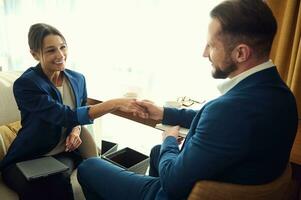 Young business partners smile at each other while shaking hands, sitting in a hotel room after successful negotiations. Young business woman at a meeting with a handsome male business partner photo