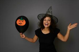 Joyful and laughing woman in magic Halloween wizard hat looks at camera, holds black balloon with felt-cut pumpkin and lifts her hand palm up, holding imaginary copy space for ad on gray background. photo