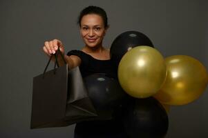 Black Friday concept with a cheerful attractive woman with beautiful smile, dressed in black holding black and golden air balloons and shopping bag, looking at camera on background with copy ad space photo