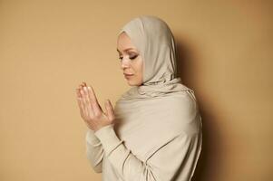 Beautiful Muslim woman wearing hijab and strict religious outfit praying on beige background with copy space. Prayer for Ramadan photo