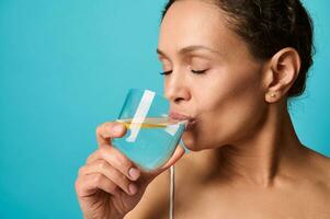 disparo a la cabeza de un hermosa africano mujer con Fresco sano piel Bebiendo limón agua desde transparente vaso, aislado terminado brillante azul antecedentes con Copiar anuncio espacio. sano comiendo y estilo de vida concepto foto