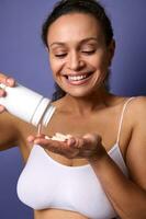 Smiling cheerful African American woman in white underwear holds a bottle of food supplement and pours a vitamin complex into her hand. Medical pills for healthy skin. Health care and medicine concept photo