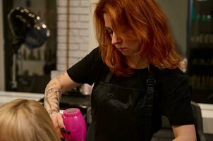 de cerca de un de edad mediana pelirrojo mujer peluquero con tatuado brazos en un estricto negro uniforme peinada, enderezamiento y el secado el rubia pelo de un cliente de un belleza salón utilizando un rosado secador de pelo foto