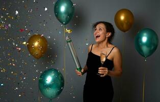 Pretty woman in evening dress holds champagne flute and throws colorful sequins confetti, celebrates Christmas, New Year, anniversary standing against gray background with inflated glitter balloons photo