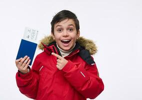 Beautiful preadolescent boy, cheerful school child in bright red warm parka points with his finger at a passport with a ticket and boarding pass, isolated over white background with copy ad space photo
