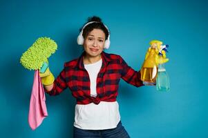 Upset woman holding cleaning supplies and looking at the camera with sadness before upcoming housework photo