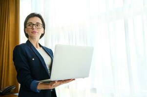 seguro de sí mismo mujer de negocios, emprendedor, periodista, ventas gerente en negocio casual traje trabajando en ordenador portátil y mirando aparte en pie por ventana en un hotel habitación. capa pluvial espacio para anuncio texto foto