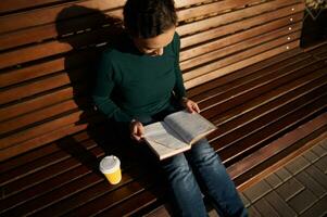 gastos generales ver de hermosa joven morena sentado en de madera banco en parque, leyendo libro y Bebiendo café. sereno confidente mujer en casual mezclilla con un libro y para llevar papel taza de caliente bebida foto