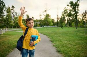 A handsome friendly charming schoolboy 9 years old, in the park, with a backpack on one shoulder and notebooks in his hands, waves his hand, looks around and smiles sweetly with a toothy smile. photo