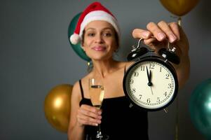 Soft focus on a black alarm clock with midnight on the clock face in the hand of a cheerful woman wearing Santa hat and holding a champagne flute on festive gray background with air balls. Christmas photo