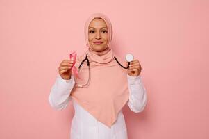 Portrait of Arab Muslim female doctor with covered head in pink hijab, posing on colored background with a pink ribbon and stethoscope in hands, supporting cancer patients and survivors, 1 st October photo