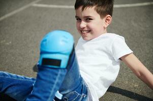 de cerca retrato de un hermosa niño en protector rodilla almohadillas sentado en el asfalto de un Deportes suelo en el calle foto