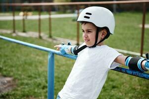 Handsome boy in safety helmet and protective gear leaning against horizontal bar on playground outdoors photo