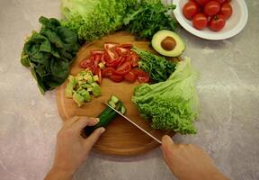 Top view of hands chopping cucumber on a wooden board next to variety of delicious healthy vegetable on the kitchen countertop. Vegan and healthy lifestyle concepts. photo