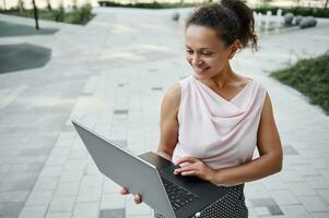 Gorgeous Hispanic young business freelancer woman cutely smiling with toothy smile while working on laptop, standing on the urban buildings background. Business and communication concept photo