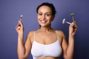 Delightful pretty woman with vitiligo skin problems smiles with beautiful toothy smile holding roller massagers near her face, confidently looking at camera posing on violet background. Copy space photo