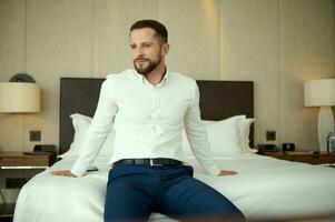 Confident portrait of a handsome middle aged bearded successful businessman, entrepreneur at business trip, thoughtfully looking aside resting, sitting on the bed in hotel room photo