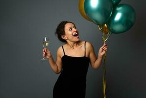 Amazed African American woman rejoices looking at festive air balls of green metallic and golden colors in her hands, holds a champagne flute isolated over gray wall background with copy space for ad photo