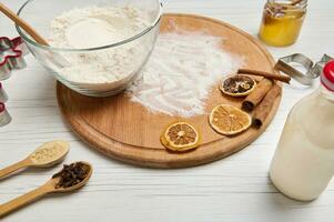 Close-up of ingredients for preparing dough and gingerbread cookies, cutting molds, bottle with plant based milk, glass jar with honey and a bowl with flour on a round wooden board photo