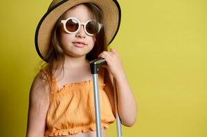 de cerca retrato pequeño niña en Gafas de sol, Paja sombrero y naranja arriba, posando con Mano equipaje, aislado en amarillo foto