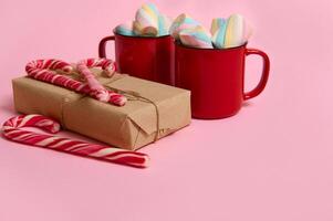 Red coffee mugs with colorful marshmallows, Christmas gift in craft wrapping paper and striped candy canes on pink colored background. Christmas and New year concept with copy space for ad photo