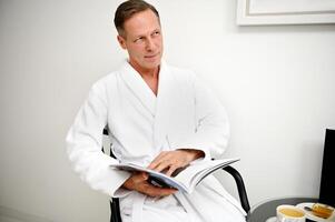 Handsome European middle aged business man wearing white terry bathrobe, sitting on an armchair and reading magazine while resting in hotel room of wellness spa center photo