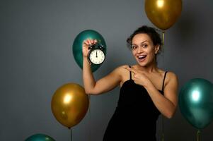 Happy woman in evening dress, rejoices at party, shows the time in the alarm clock in her hand, it's midnight. Celebrating Christmas, New Year, birthday concept on gray background with copy space photo