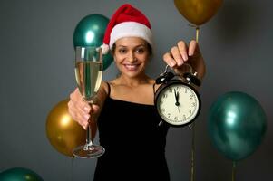 Focus on alarm clock with midnight on the clock face and sparkling wine glass in the hands of cheerful woman wearing Santa hat and smiling festive gray background with shiny air balls for Christmas ad photo
