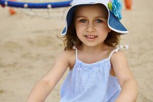 Face portrait of a cute beautiful little girl wearing sunhat and relaxing on the sand background. Summer camp photo