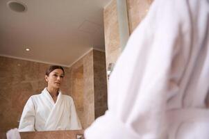 Mirror reflection of an attractive confident middle aged brunette woman wearing bathrobe looking at herself standing in bathroom during morning hygiene routine photo