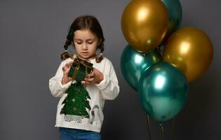 Adorable girl holds her Christmas gift in green glitter wrapping paper and golden ribbon, stands near beautiful shiny green metallic and golden air balloons , isolated over gray background, copy space photo