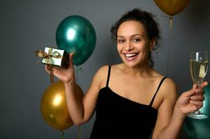 Smiling mixed race woman with beautiful toothy smile rejoicing, posing with a Christmas gift and flute of sparkling wine in her hands, against air balloons on gray background. New Year concept for ad photo