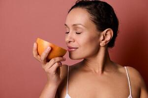 Close-up of dark-haired pretty woman smelling the scent of fresh juicy red grapefruit, posing with closed eyes against coral background with copy space. Health, body positivity and skin care concept. photo