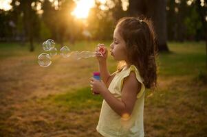 Adorable child baby girl, blowing soap bubbles at sunset, enjoying pleasant time outdoors in the meadow. Sun's rays fall through transparent bubble spheres with iridescent reflections. photo