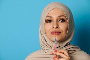 Closeup portrait of pretty Muslim woman with covered head in hijab holding a syringe with beauty injection near her lips. photo